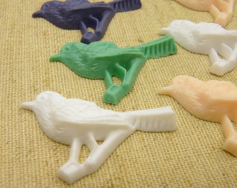 May include: Five small bird figurines made of different coloured plastic. The birds are arranged in a row on a brown hessian surface. The birds are all facing the right side of the image. The birds are all different colours: purple, green, white, peach, and grey.