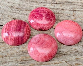 May include: Four pink and white cabochon stones with a polished finish. The stones have a striated pattern and are arranged on a wooden surface.