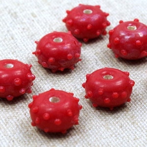May include: Seven red, round beads with a textured surface and a hole in the centre. The beads are arranged in a semi-circle on a light brown fabric surface.