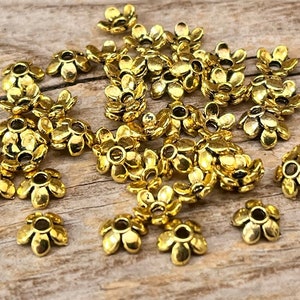 Puede incluir: Un grupo de tapas de cuentas de metal de color dorado en forma de flor. Las tapas tienen un pequeño agujero en el centro para enhebrar.