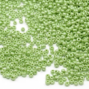Può includere: Una grande quantità di piccole perline rotonde di vetro verde. Le perline hanno una finitura perlescente e sono sparse su una superficie bianca.