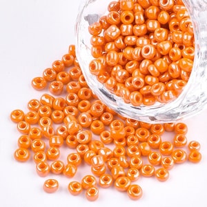 Può includere: Una grande quantità di piccole perline di vetro rotonde di colore arancione. Le perline sono sparse su una superficie bianca e fuoriescono da una ciotola di vetro trasparente.