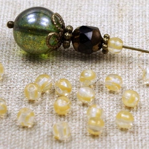 Könnte beinhalten: Eine dekorative Hutnadel mit einer grünen und goldenen Glaskugel, einer facettierten dunkelbraunen Perle und kleinen gelben Perlen. Verstreut sind kleine, durchscheinende gelbe Perlen mit weißen Wirbeln. Die Nadel befindet sich auf einer strukturierten, neutralfarbenen Oberfläche.