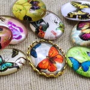 Könnte beinhalten: Sortiment ovaler Glascabochons mit Schmetterlingsmotiven. Die Cabochons sind in verschiedenen Farben gehalten, darunter Rosa, Blau, Grün, Gelb und Weiß. Einige Cabochons haben eine Vintage-Ästhetik mit Blumenmustern und Text.