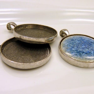 Könnte beinhalten: Zwei silberfarbene Anhänger-Tabletts mit einer strukturierten Oberfläche und einer Öse zum Aufhängen. Ein Tablett ist mit einem blauen und weißen glitzernden Material gefüllt.