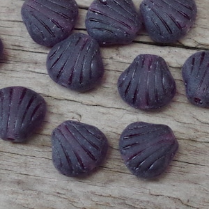Könnte beinhalten: Eine Sammlung von lila Glasperlen in Form von Muscheln. Die Perlen haben eine strukturierte Oberfläche und sind auf einer Holzoberfläche angeordnet.