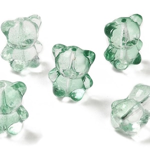 Könnte beinhalten: Fünf transparente grüne Glasperlen in Bärenform. Die Perlen sind durchscheinend und haben ein Loch in der Mitte zum Aufziehen.