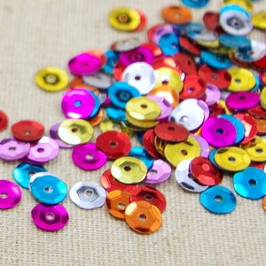Könnte beinhalten: Ein Haufen bunter, schimmernder Pailletten in verschiedenen Farbtönen von Rot, Rosa, Blau, Gelb und Silber. Die Pailletten sind auf einer beigefarbenen Stoffoberfläche verstreut.