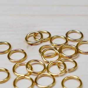 Könnte beinhalten: Eine Gruppe goldener Sprungringe, auch bekannt als Splintringe, liegt auf einer hellbraunen Holzoberfläche verstreut. Diese Ringe werden zum Verbinden von Schmuckkomponenten verwendet.