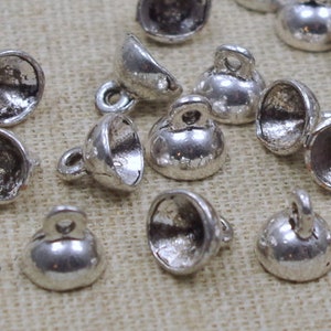 Puede incluir: Una colección de cuentas de metal de color plateado en forma de cúpula con un bucle en la parte superior. Las cuentas están esparcidas sobre una superficie de tela texturizada.