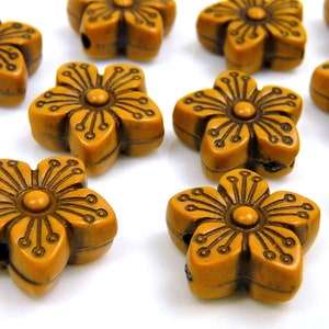 May include: Close-up of several flower-shaped beads. The beads are a warm, golden-brown color with darker brown etched details. Each bead has a central circle and five petals, with small holes for stringing.