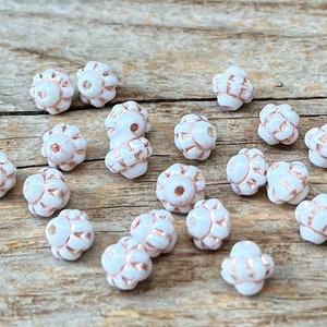 Könnte beinhalten: Eine Sammlung weißer Glasperlen mit einer roségoldenen Metallic-Oberfläche. Die Perlen haben ein blumenartiges Design und sind perfekt für die Schmuckherstellung.