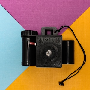 Può includere: Una macchina fotografica giocattolo in plastica nera con un obiettivo rosso e un cinturino nero. La macchina fotografica ha la scritta "SNAPSHOOTER" stampata sul davanti.