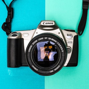 May include: A silver and black Canon EOS Rebel camera with a black lens and strap. The lens reads "CANON ZOOM LENS EF 28-80mm 1:3.5-5.6 II". The camera is set against a blue and teal background.
