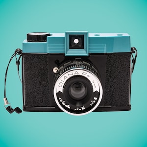 Puede incluir: Una cámara Diana F+ azul y negra con una lente de 75 mm. La cámara tiene una correa negra y una tapa de lente plateada.