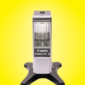 May include: A Canon Canolite D flash unit, silver and black, is positioned on a black base against a yellow backdrop. The flash has a rectangular form with a grid-like front and the Canon logo.