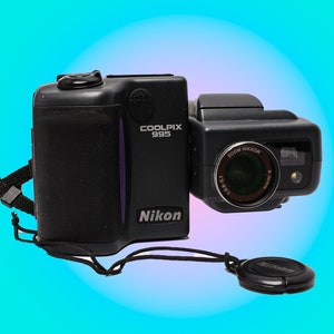 May include: Black Nikon Coolpix 995 digital camera with a Zoom Nikkor lens. The camera has a lens cap attached and a black strap. The camera is on a gradient blue and pink background.