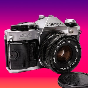 Puede incluir: Una cámara de película Canon AE-1 Program plateada con una lente Canon FD 50mm 1:1.8. La cámara tiene una cubierta de imitación de cuero negro y una tapa de lente negra.