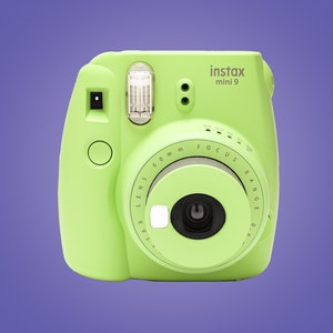 Peut inclure: Un appareil photo instantané Instax Mini 9 vert clair. L'appareil photo dispose d'un flash intégré, d'un objectif avec une plage de mise au point de 60 mm et d'un viseur. L'appareil photo est sur un fond violet.