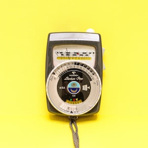 May include: A vintage Gossen Luna-Pro light meter with a black and silver body. The meter has a dial with numbers and markings, including ASA, time, and f-stops. A small cord hangs from the bottom. The background is a solid yellow.