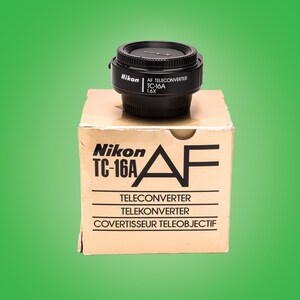 Könnte beinhalten: Ein schwarzes Nikon TC-16A AF Telekonverter-Objektiv in seiner Originalverpackung. Die Schachtel ist braun mit schwarzem Text, der "Nikon TC-16A AF Teleconverter Telekonverter Covertisseur Teleobjectif" lautet.