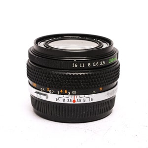 May include: A black 28mm camera lens with a silver ring and markings for aperture and distance in both feet and meters. The lens has a black rubber ring around the front element.