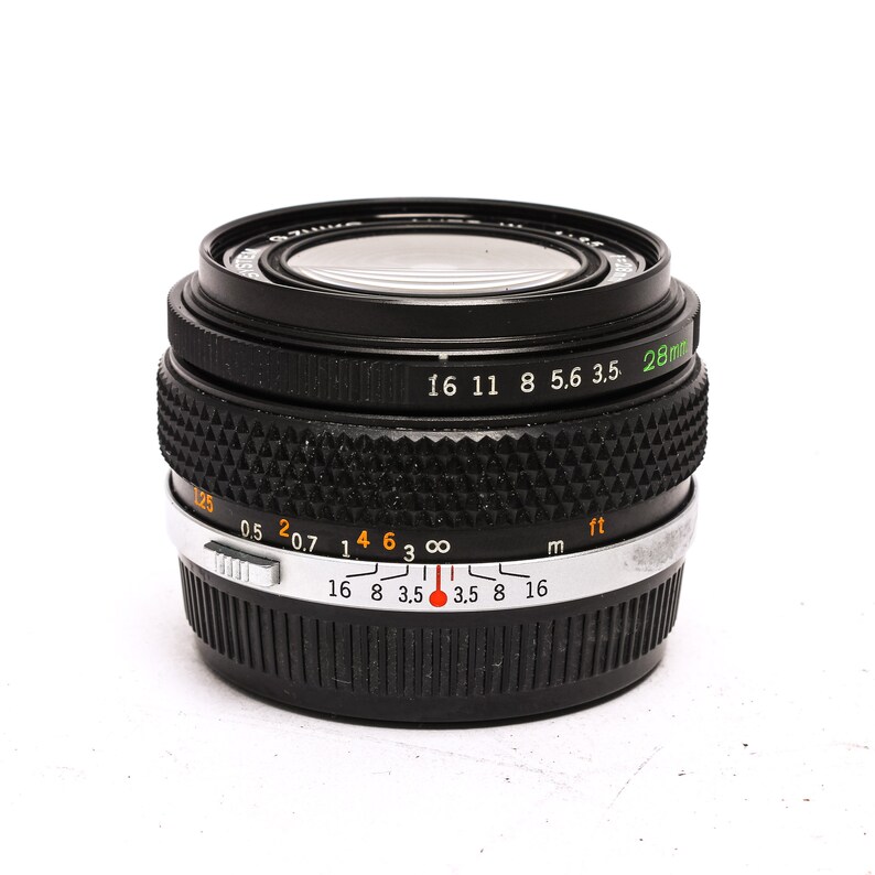 May include: A black 28mm camera lens with a silver ring and markings for aperture and distance in both feet and meters. The lens has a black rubber ring around the front element.