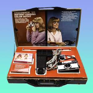 May include: An open box containing an Anscomatic 126 instant loading colour outfit. The box includes a camera, projector, film, and instructions. The camera is black with a silver lens and a red button. The projector is black with a silver lens and a red button. The film is in a clear plastic case. The instructions are in a white booklet.