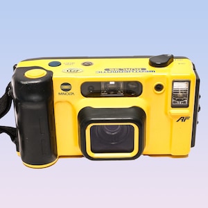 May include: A yellow Minolta Weathermatic Dual 35 film camera with a black grip and lens. The camera has a black autofocus button and a black flash. The camera has a black and white logo that says "Minolta" and the text "Weathermatic Dual 35" on the top.