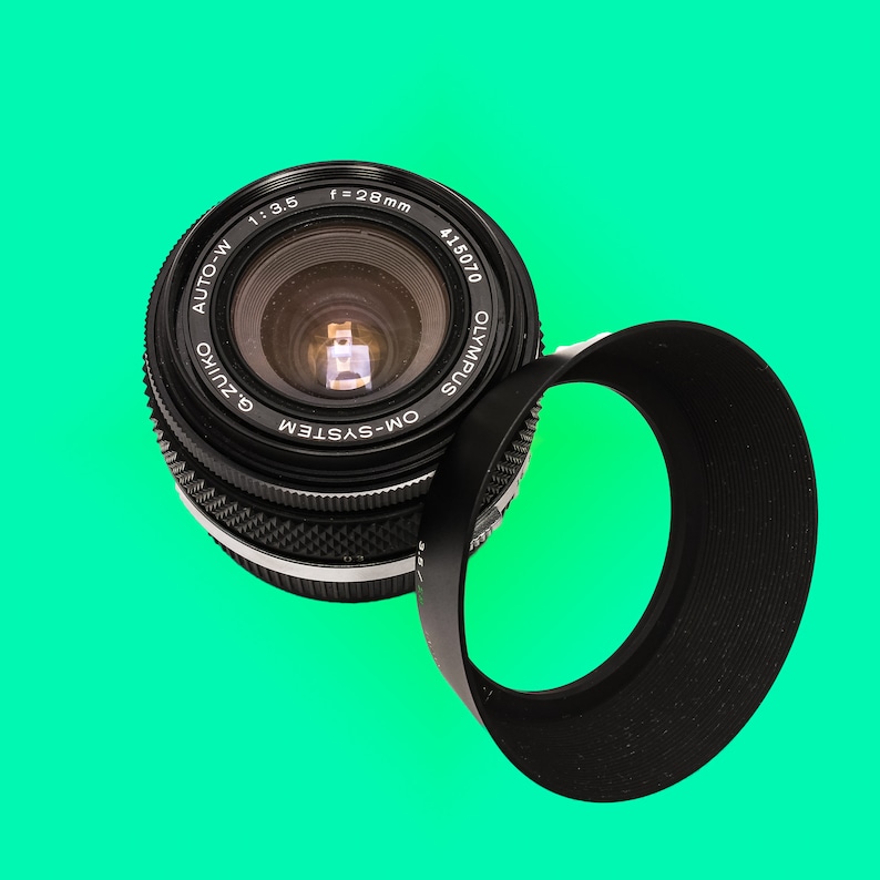 May include: A black camera lens with a silver ring around the edge. The lens has the text "OM-SYSTEM G.ZUIKO AUTO-W 1:3.5 f=28mm 415070 OLYMPUS" printed on it. A black lens hood is attached to the lens.