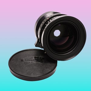 Op de afbeelding: Een zwarte cameralens met een lensdop. De lens heeft meetmarkeringen en een groot frontelement. De lensdop is zwart en heeft de tekst "SCHNEIDER-KREUZNACH" erop gedrukt. De achtergrond is een verloop van roze en blauw.