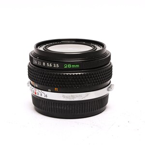 May include: A black 28mm camera lens with a silver ring and markings for aperture and distance. The lens has a black rubber ring around the front element.