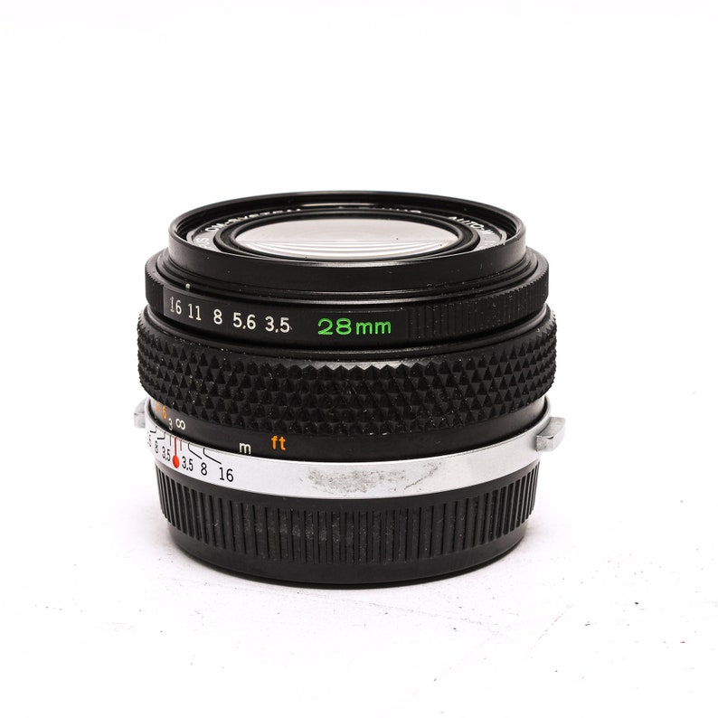May include: A black 28mm camera lens with a silver ring and markings for aperture and distance. The lens has a black rubber ring around the front element.