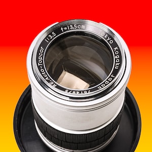 Può includere: Un obiettivo per fotocamera argentato con dettagli neri. L'obiettivo ha il testo "Tokyo Kogaku Japan 7614875 RE, Auto-Topcor 1:3.5 f=13.5cm" stampato attorno al bordo.