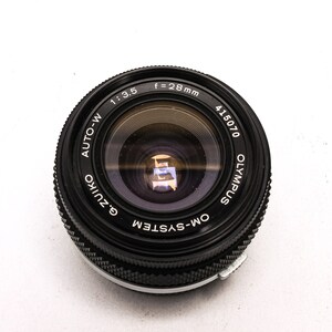 May include: A black Olympus OM-System Zuiko Auto-W 28mm f/3.5 lens with a silver ring around the edge. The lens has a black lens cap and the text "Olympus", "Zuiko", "Auto-W", "OM-System", "f=28mm", "1:3.5", and "415070" is printed on the lens.