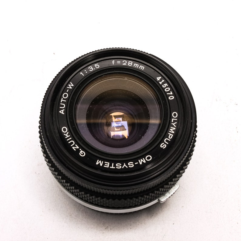 May include: A black Olympus OM-System Zuiko Auto-W 28mm f/3.5 lens with a silver ring around the edge. The lens has a black lens cap and the text "Olympus", "Zuiko", "Auto-W", "OM-System", "f=28mm", "1:3.5", and "415070" is printed on the lens.