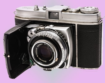 May include: A silver and black 35mm film camera with a Schneider-Kreuznach 28mm lens. The camera has a built-in light meter and a rangefinder. The camera is in good condition and has a few signs of wear.