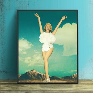 May include: A woman in a white swimsuit stands with her arms raised in the air, smiling and laughing. She is surrounded by a large white cloud, resembling a mushroom cloud, against a blue sky with white clouds. The woman is standing on a brown, rocky surface.