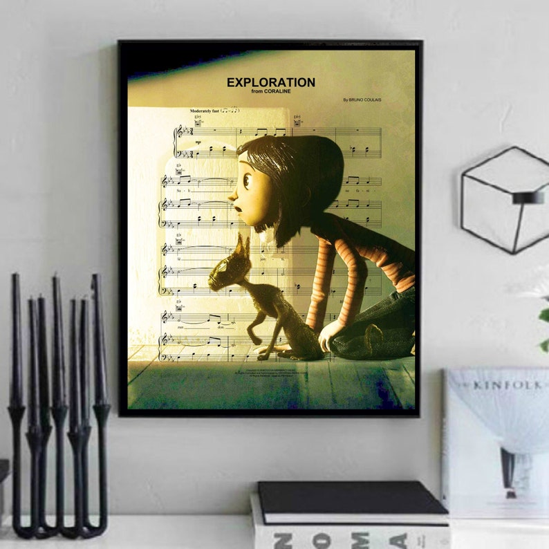 May include: A framed print of a musical score with the title "Exploration from Coraline" by Bruno Coulais. The score features a girl with brown hair and a red and white striped shirt looking at a cat with green eyes. The image is set against a light yellow background.