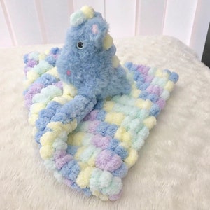 May include: A blue and white plush toy dinosaur with a light blue pom-pom blanket. The blanket is made of soft yarn and has a checkered pattern.
