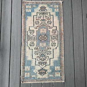 May include: A rectangular, vintage-style rug with a central geometric design. The rug features a light blue border and a cream-colored central field with intricate patterns in shades of blue, brown, and beige. The rug is likely made of woven material.