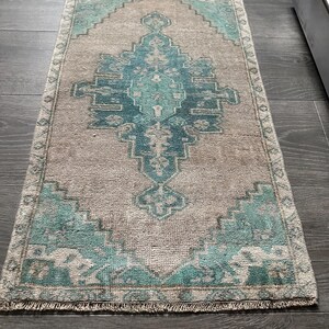 May include: A rectangular rug with a distressed appearance, featuring a central geometric design in teal and beige. The rug has a beige backdrop with teal detailing and a fringed border. It rests on a dark wooden floor.