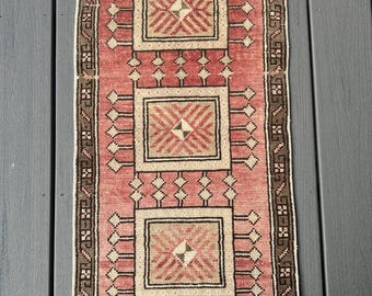 Op de afbeelding: Een rechthoekig, handgeweven tapijt met een centraal ontwerp van drie beige vierkanten, elk met een geometrisch patroon. Het tapijt heeft een roze achtergrond en een bruine rand met een herhalend patroon. Het tapijt is ongeveer 91 cm lang en 51 cm breed.