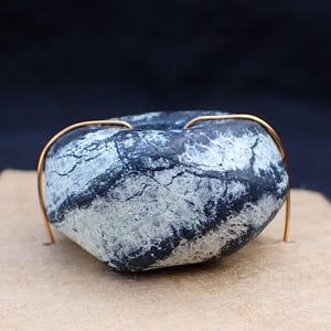 May include: A heart-shaped stone with a gold wire wrap, resting on a brown felt square. The stone has a dark blue and white marbled pattern. The background is a dark blue.
