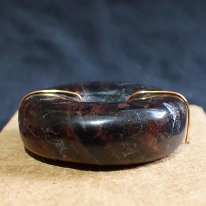May include: A dark, ring-shaped object with a polished, speckled surface, possibly stone or a similar material. Thin, gold-colored wire accents the top. The object rests on a folded piece of brown paper, set against a dark background.
