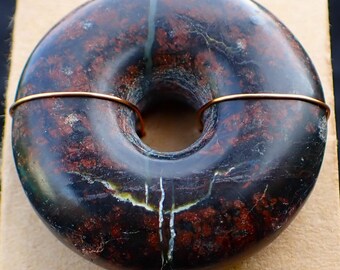 Puede incluir: Un colgante de piedra oscura en forma de rosquilla con vetas rojas y negras, atravesado por un fino alambre de cobre. La piedra descansa sobre un trozo de papel rectangular de color beige, sobre un fondo oscuro. El colgante tiene un agujero en el centro.