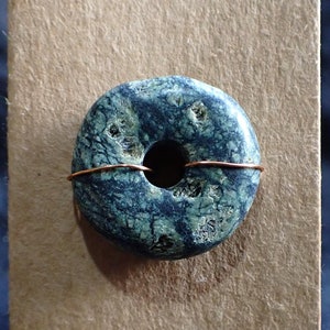 May include: A dark blue and green stone donut with a hole in the center. The stone is wrapped with a thin copper wire.