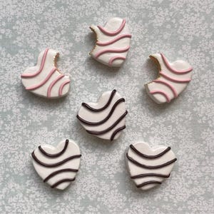 Puede incluir: Vista cenital de seis galletas en forma de corazón. Algunas son blancas con rayas rosas, otras blancas con rayas marrones. A dos de las galletas con rayas rosas les falta un mordisco. Sobre un fondo azul claro.