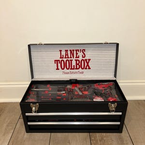 Puede incluir: Caja de herramientas negra con herramientas rojas y negras en el interior. La caja de herramientas tiene dos cajones y una tapa con bisagras. El texto "LANE'S TOOLBOX" está impreso en rojo en el interior de la tapa.