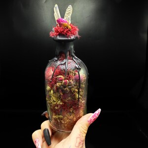 May include: A clear glass bottle filled with dried flowers and moss, topped with a cluster of crystals and a pink rose. The bottle's neck is adorned with black wax drips, creating a unique, decorative piece.